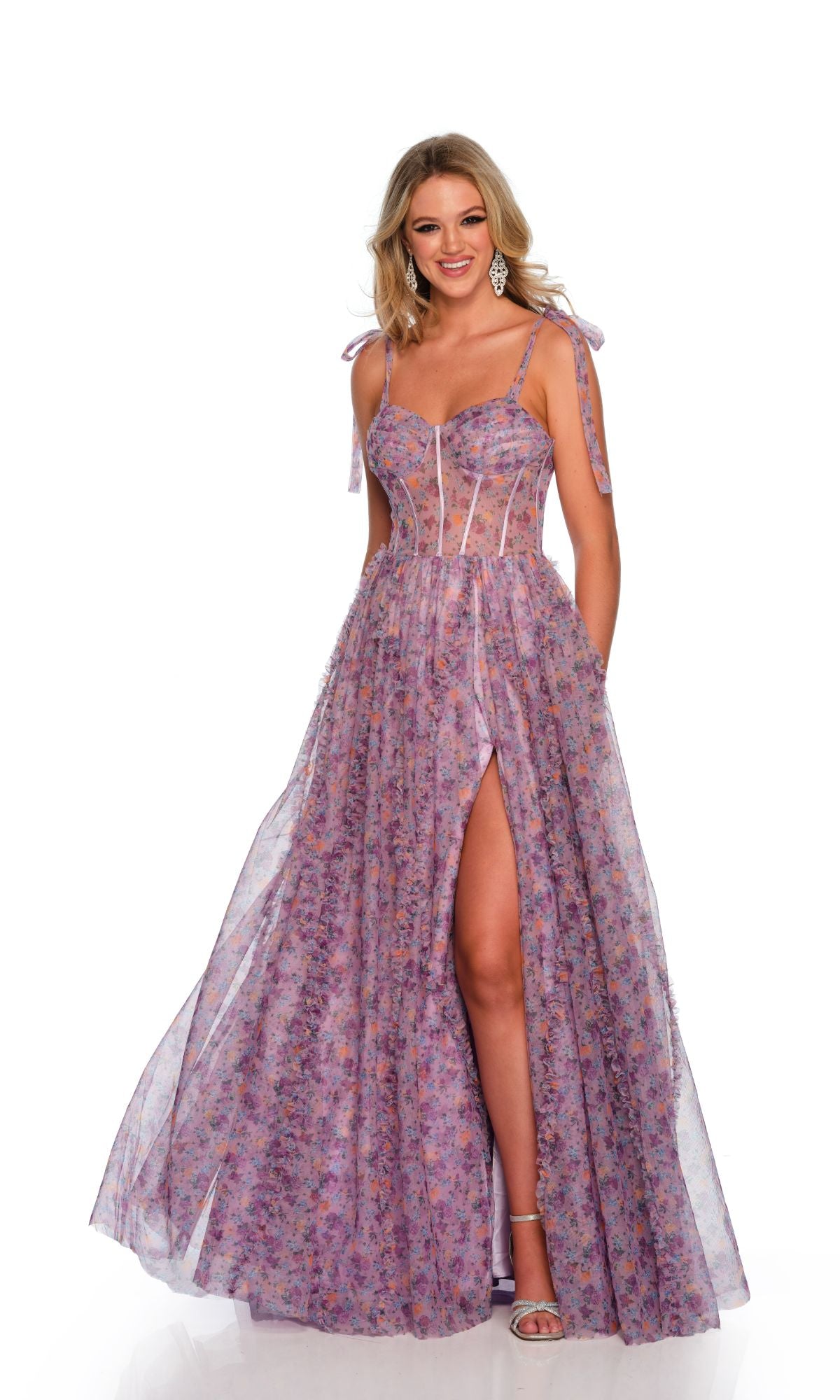 A woman wearing the Long Formal Dress 11591 by Dave and Johnny—floor-length, sleeveless, purple floral with sheer panels, sweetheart neckline, tied straps, thigh-high slit, and open lace-up back—stands against a white background.